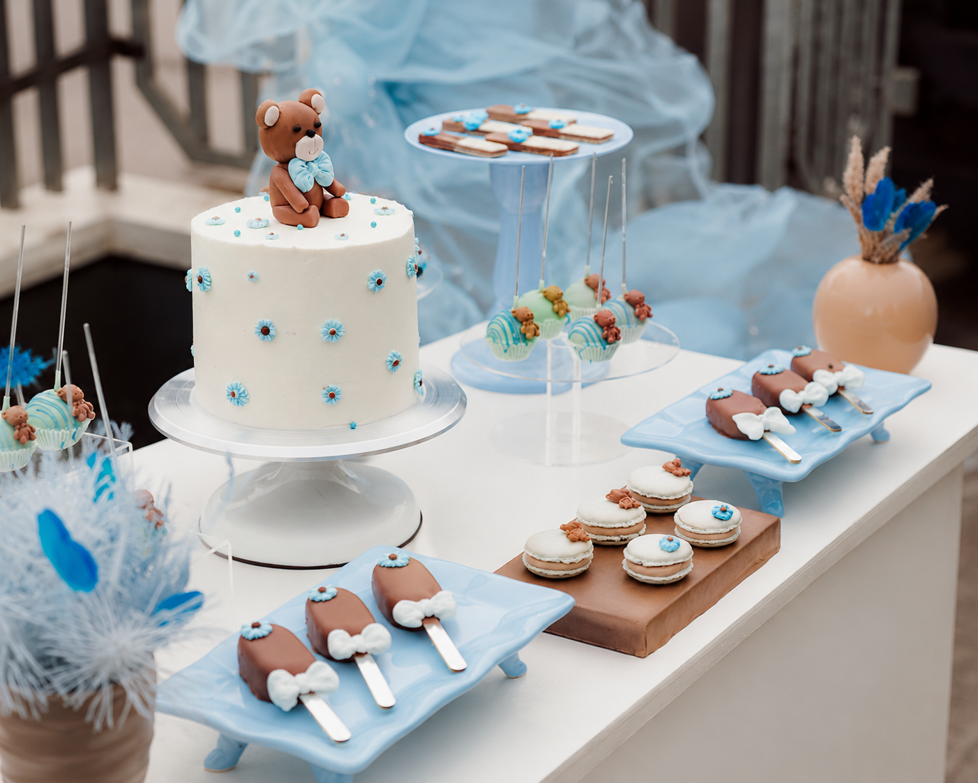 Masă elegantă cu torturi personalizate pentru evenimente, decorată cu un tort tematic cu ursuleț și flori albastre, macarons, cake pops și deserturi artizanale, într-o paletă rafinată de tonuri pastelate. Creație semnată Ady Cake Lab – site web adycakelab.com.